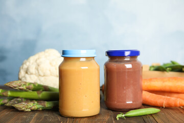 Jars with baby food and fresh vegetables on wooden table against light blue background