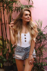 Obraz na płótnie Canvas Stylish yooung blond woman wearing white shirt and jeans posing on the background of pink wall with ropical plants