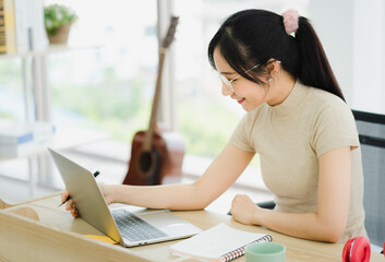 Young Asian woman in casual outfit wear glasses working at home with smile and happy emotion sitting in work desk using laptop and writing to take note in modern room