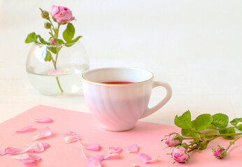 Obraz na płótnie Canvas Petals and a branch of a pale pink rose on the background of a white Cup of tea and a bouquet of flowers, close-up, place for the inscription, side view - the concept of a festive day