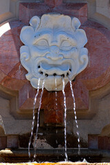 Close up image of a water fountain, where water comes as five streams through the holes located at the mouth of a stone statue. Head only statue is placed on an elevated pink platform.