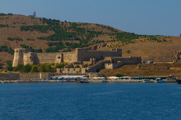 Landscape view of the coast of Bozcaada (Tenedos), Turkey. Image features a medieval castle, tourist shops, beach umbrellas and some boats along the shore.