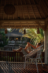 cucasian woman sit  on wicker chair at home terrace outdoor, view on the rice field. Travel blogger girl enjoy vacation in autentic Bali village
