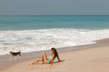 Fototapeta na wymiar Summer lifestyle portrait of pretty young suntanned woman sitting on the beach of the tropical island and look at the dog walk next to her