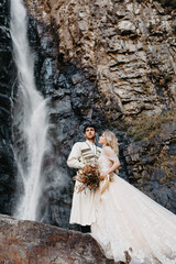 noise effect, selective focus: incredibly enamored brides hugging, kissing and posing for a photo on the incredible rocky mountains background with a big waterfall