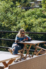 photo through window: stylish attractive girl dressed in warm clothes sits on the terrace of a mountain cafe and lonely smokes a cigarette in warm sunny weather