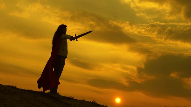 girl plays roman lenin in bright rays of sun against sky. free woman knight. super woman with a sword in his hand and in red cloak stands on mountain in sunset light. free woman playing superhero.