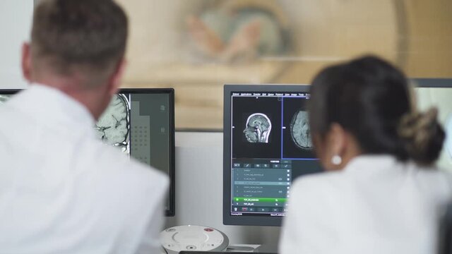Magnetic resonance imaging, adult man and asian woman professional doctors examines the results of magnetic resonance therapy of brain, looks at screens.