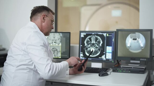 Magnetic resonance imaging, adult professional doctor examines the results of magnetic resonance therapy of brain, looks at screens and uses a screen tablet.
