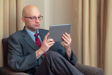 Bald attractive manager businessmen sitting on a chair in his hotel room looking at tablet. Man in grey suit, blue shirt, dark red tie, and metal glasses.