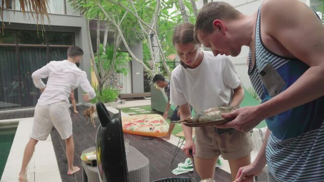 Handheld shot of young man and woman cooking fish barbecue while their friends laying frisbee in Bailan villa yard