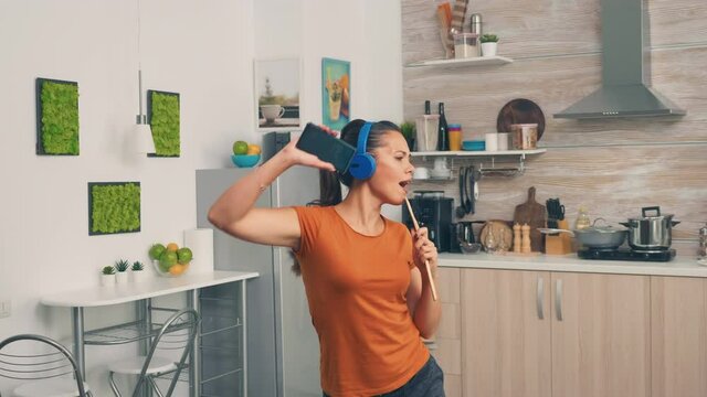 Positive young woman singing on wooden spoon in the morning. Energetic, positive, happy, funny and cute housewife dancing alone in the house. Entertainment and leiuse alone at home