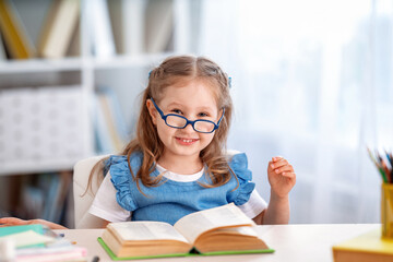 happy smart little girl with glasses is sitting at a table with a book in room.