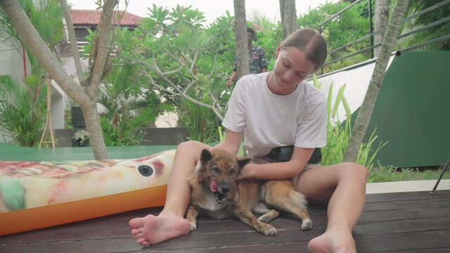 Joyful Caucasian girl wearing white T-shirt and shorts sitting outdoors petting her lovely dog, young man playing frisbee behind them