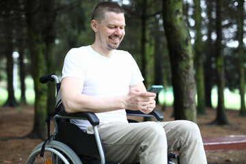 Portrait of cheerful happy person sitting in wheelchair and using smartphone for fun. Laughing man in white shirt having fun in forest. Smiling adult enjoying life on nature. Disabled people concept