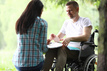 Fototapeta na wymiar Portrait of smiling man sitting in wheelchair with papers. Handsome middle aged person in white shirt. Woman holding folder with documents. Working moments. Business and disabled people concept