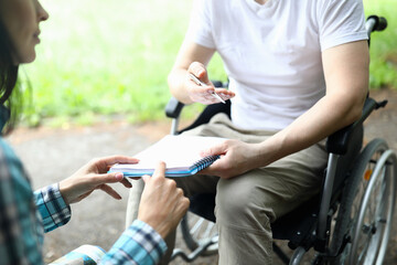 Close-up of man sitting in wheelchair and holding paper for signing. Woman showing place for signature. Business moment and meeting in park. Disabled people concept