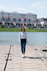 Smiling, pretty girl with long hair in a white blouse goes on the banks of the river city, against the backdrop of new homes