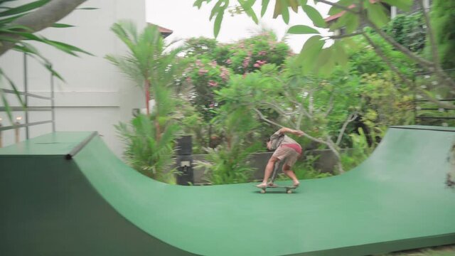Handheld shot footage of active young man riding skateboard on half pipe in Bali villa yard loosing balance and falling down