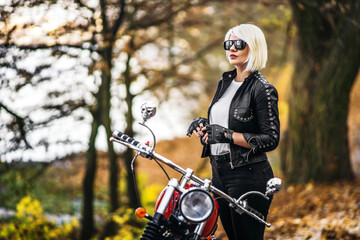 Pretty blonde biker girl in sunglasses with red motorcycle on the road in the forest