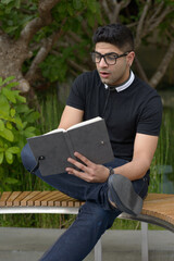 Fototapeta premium Young handsome Indian man with eyeglasses reading book and looking shocked at the park
