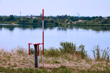 Fototapeta na wymiar Well with a measuring bar for monitoring the groundwater level and water quality next to a gravel pond