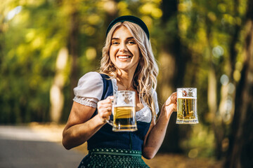 Pretty happy blonde in dirndl, traditional festival dress, holding two mugs of beer outdoors in the...