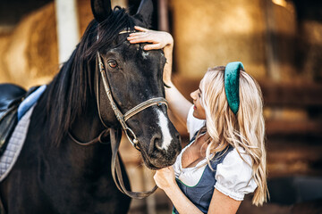 Obraz premium Pretty blonde in traditional dress take care of big black horse at the farm