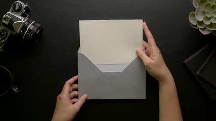 Female hands open mock-up greeting card with grey envelope on dark office desk with camera and decorations