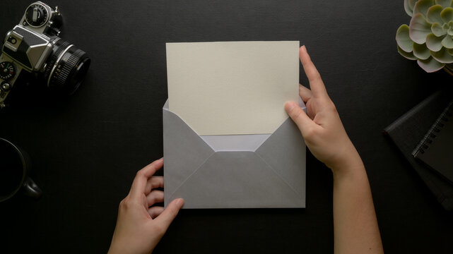 Female hands open mock-up greeting card with grey envelope on dark office desk with camera and decorations