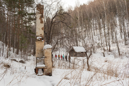 Old chimney. Mountain valley in winter, white snow, trees. People walk along the trail. Concept of an abandoned settlement, expedition, tourism.