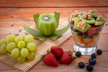A glass cup with a fresh and colorful fruit salad ready to eat. Some fruits on the table made of recycled wood