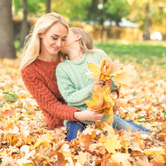 Happy young caucasian mother and little daughter holding autumn yellow leaves sitting and kissing at the park