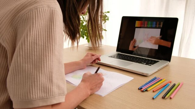 Side view of crop anonymous female artist drawing on paper sheet with multicolored pencils while sitting at wooden desk in front of open netbook with video instruction