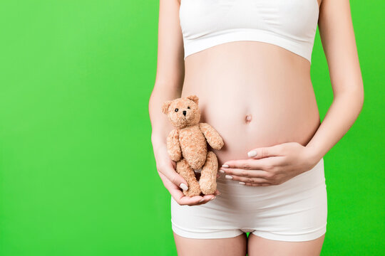 Cropped image of happy pregnant woman in white underwear holding teddy bear against her belly at green background. Child expecting. Copy space