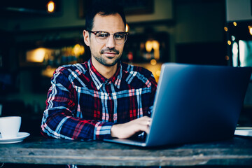 Portrait of smiling successful young man in optical eyeglasses updating software on digital laptop device connected to wireless 4G internet while looking at camera sitting in stylish coffee shop