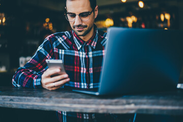 Cropped image of bearded student in optical spectacles reading blog on telephone device while preparing for seminar sitting at laptop computer for searching new information for productive work