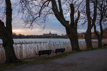 Empty park benches and leafless trees along the Drugardsbrunnsviken waterway at a winter sunset.