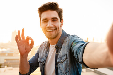 Photo of joyful young man gesturing ok sign while taking selfie