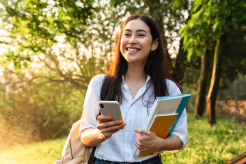 Photo of happy asian student woman using cellphone while walking in parkPhoto of happy asian student woman with exercise books using cellphone while walking in green park