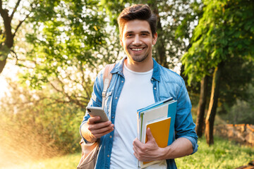 Photo of smiling student man using smartphone while walking in park