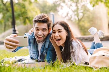 Image of happy couple taking selfie on smartphone while lying on grass