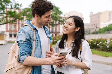 Image of joyful student couple talking and using cellphone while walking