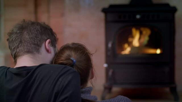 Happy loving couple sitting near fireplace in country house hugging, young woman puts her head on man's shoulder, close-up, back view. Warm romantic evening, relationship, love, leisure time