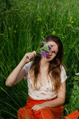 Young brunette girl in white top and red skirt sitting in green field grass in countryside smelling small bouquet of field flowers in hands with closed eyes and enjoy nature and rural life.