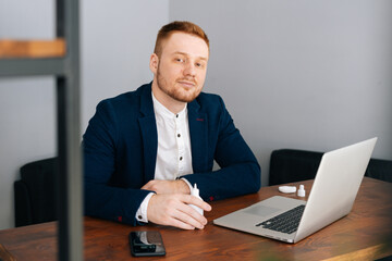 Skilled young businessman wearing fashion suit is applying sanitizer to hands sitting at the desk in modern office room ,looking at the camera.. Concept of office working.