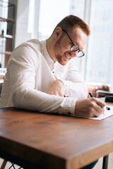Fototapeta na wymiar Cheerful young man wearing fashion casual clothing and stylish eyeglasses is writing text on paper while sitting at the wooden desk in modern room on background of large window in sunny day.