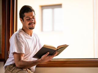 A picture of a brunette male sitting next to the window and reading a book