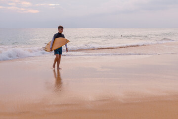 Handsome teenage boy with surfboard walking along the ocean beach at  sandy coast.