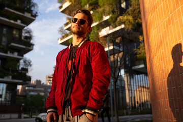 The front portrait of the young man with sunglasses and a red jacket, looking confident at camera of the urban street in the city of Milan being sunset.